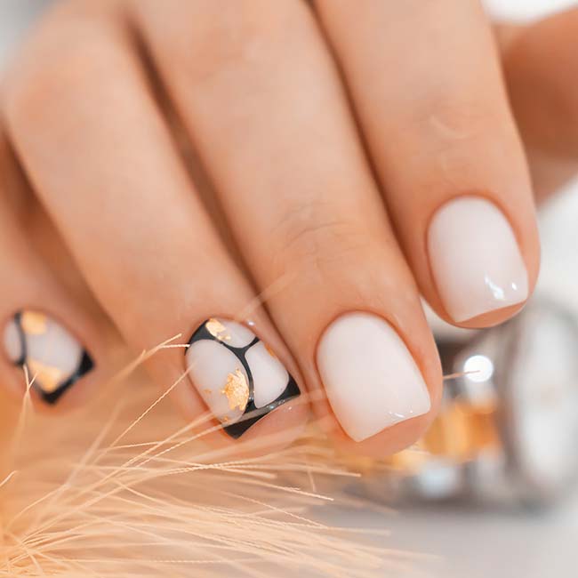 Hand with short nails, white polish, abstract black and gold leaf design, and nail enhancements.