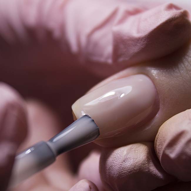 A gloved hand from The Nail Department and More applies clear nail polish to rebalance nails.