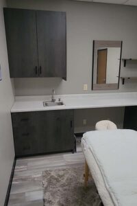 A massage room with a sink, cabinets, mirror, and skincare essentials on the white-sheeted table.