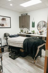A cozy spa room with a massage table, blankets, skincare decor, and a round mirror.