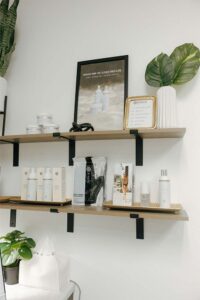 Two wooden shelves display skincare and natural nail care products, plants, and framed signs.
