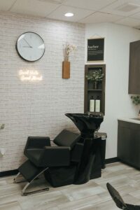 A black salon shampoo chair under a clock and neon at The Nail Department and More.