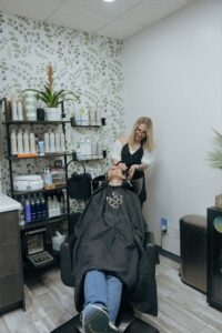 A hairstylist washes a client's hair at The Nail Department and More, beside product shelves.