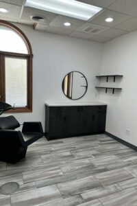 Modern salon room for Nail Enhancements with a black chair, round mirror, and gray tile floor.