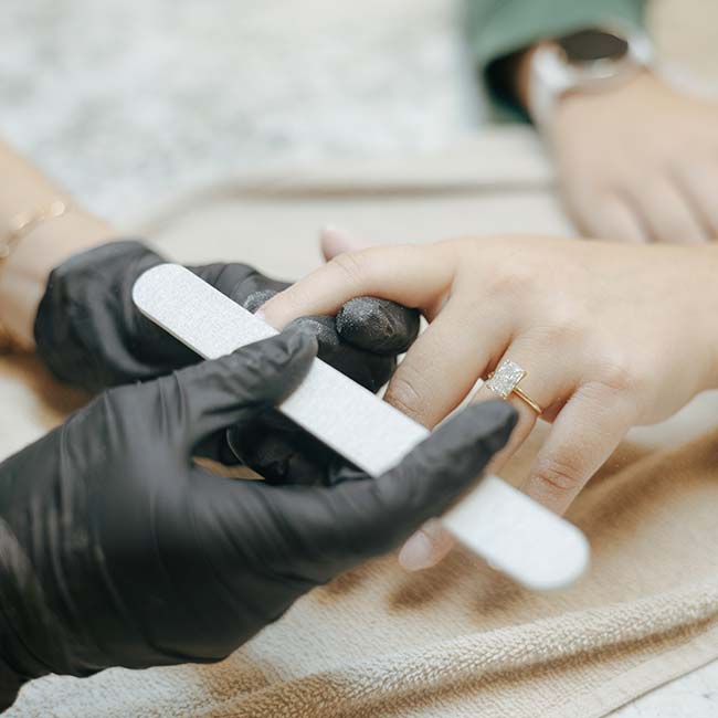 A gloved cosmetologist files a woman's nails; she wears a gold ring with a large stone.
