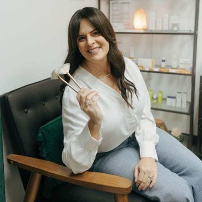 Woman in white blouse and jeans sits, smiling and holding facial rollers for skincare rebalance.