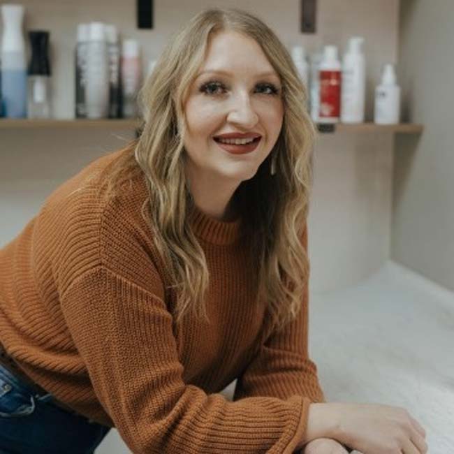 Smiling woman with wavy hair, a brown sweater, and shelves of hair products for cosmetologists.
