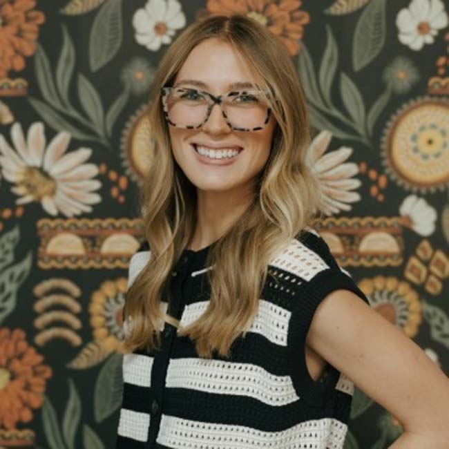 Smiling woman with glasses and striped top poses by a floral wall at The Nail Department and More.