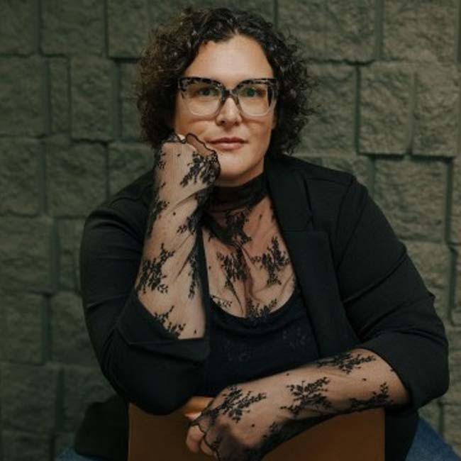 Woman with curly hair and glasses, stylish at The Nail Department and More, sits thoughtfully.