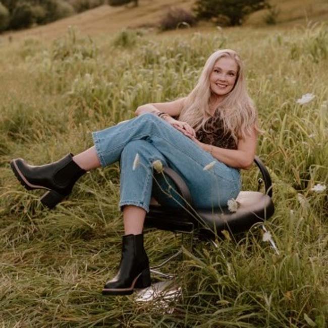 Woman with long blonde hair sits on a chair in a grassy field, smiling at The Nail Department and More.