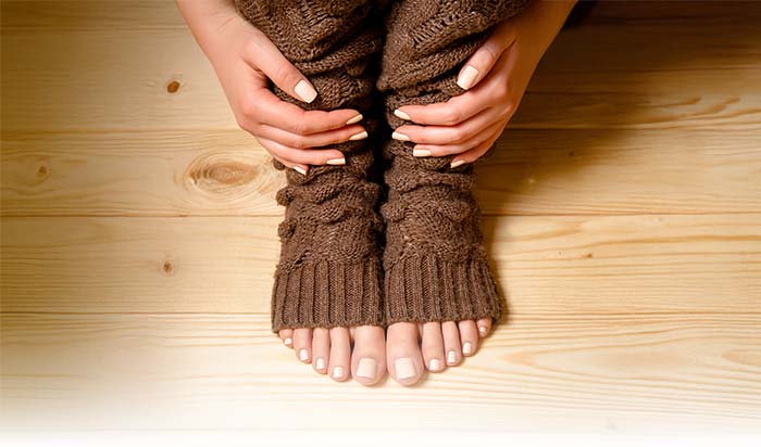 Hands resting on knees with brown leg warmers—bare feet showing natural nail care on wood floor.