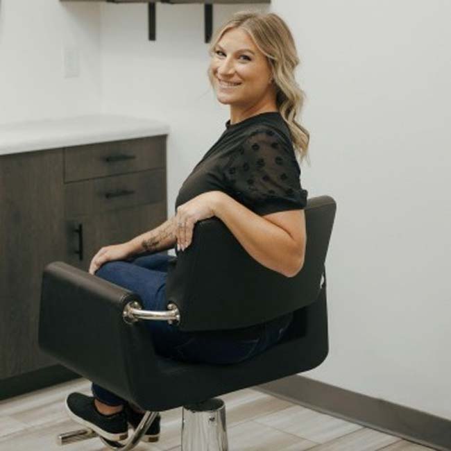 Smiling woman with blonde hair sits sideways in a salon chair, showcasing natural nail care.