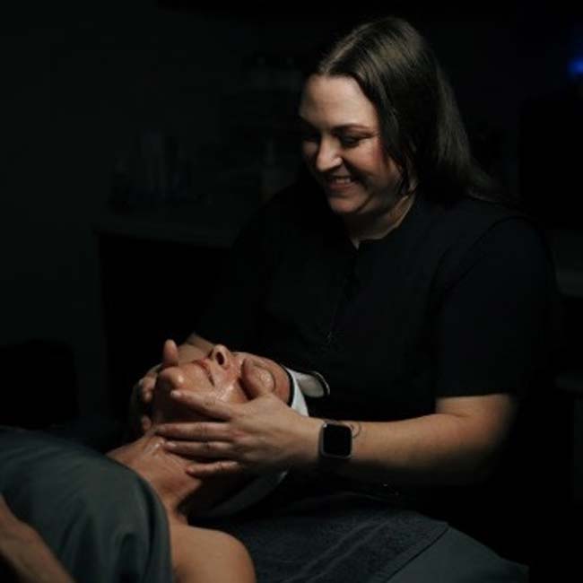 A cosmetologist smiles while giving a facial massage in The Nail Department and More's dim room.