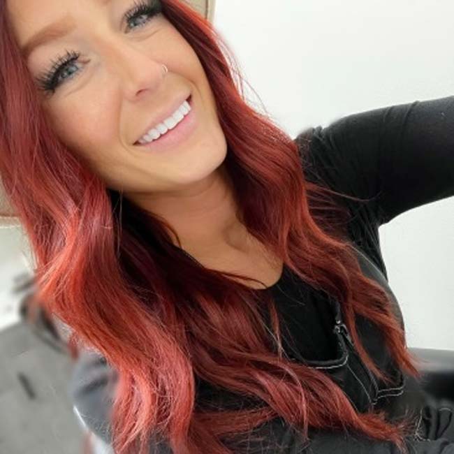 Smiling woman with long wavy red hair, flaunting skincare glow while taking a selfie in a beige hat.